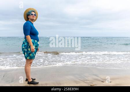 Un uomo transgender, si trova su una spiaggia che guarda verso il mare. Indossando un top blu, una gonna floreale e un cappello di paglia, mostra un comportamento pacifico in mezzo a questo Foto Stock