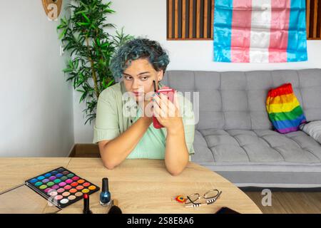 Un giovane uomo transgender applica con cura il trucco, seduto a un tavolo con vari oggetti per il trucco. Sullo sfondo è visibile una bandiera di orgoglio, Symboliz Foto Stock