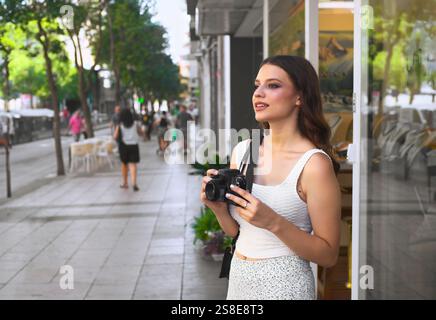 Una giovane donna si trova in una vivace strada della città, tenendo una macchina fotografica circondata dai passanti, ammira il paesaggio urbano gli alberi e i negozi fiancheggiano il liv Foto Stock