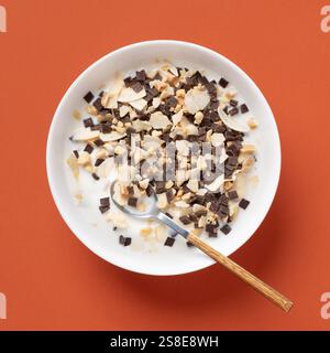 Vista dall'alto di una ciotola bianca piena di cereali, mandorle affettate e pezzi di cioccolato, tutto immerso nel latte, su un vivace sfondo arancione. A W Foto Stock
