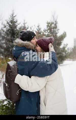 Un uomo bacia amorevolmente una donna sulla fronte mentre si abbracciano in un parco innevato vestito con cappotti e cappelli invernali, condividono un momento tenero circondato Foto Stock