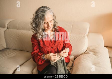 Una donna dai capelli grigi si siede comodamente su un divano beige, concentrandosi sulla lavorazione a maglia. Indossa una camicetta rossa, trasmettendo calore e creatività in un ambiente accogliente Foto Stock