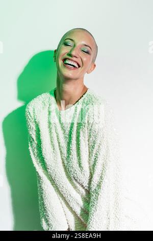 Una donna allegra di mezza età con la testa rasata sorride brillantemente. Indossa un maglione bianco testurizzato, in piedi su uno sfondo chiaro con li verde Foto Stock