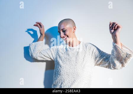 Una donna di mezza età con la testa rasata si posa gioiosamente su uno sfondo neutro, celebrando l'individualità e la fiducia in un radioso sudore bianco Foto Stock