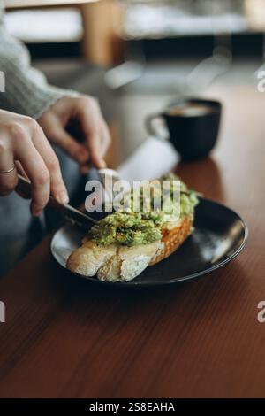 Un ambiente accogliente per la colazione con una persona che prepara un brindisi all'avocado su un piatto scuro, accompagnato da una tazza di caffè nero, sistemato su un tavolo di legno in un bar Foto Stock
