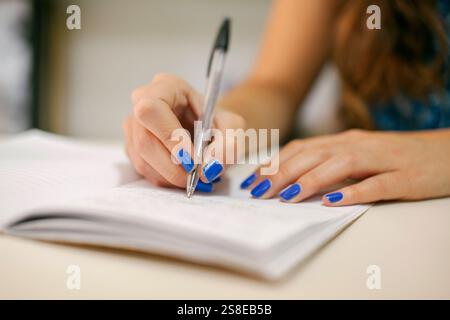 Primo piano di una mano con unghie dipinte in blu che scrivono su un taccuino. L'attenzione è sulla penna e sulla mano, sottolineando la creatività, la pianificazione e l'organizzazione Foto Stock