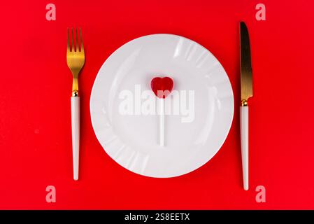 Posate di San Valentino con piatto, coltello, forchetta e lollypop a forma di cuore di zucchero rosso, su sfondo rosso brillante, vista dall'alto spazio di copia. Festività, va Foto Stock