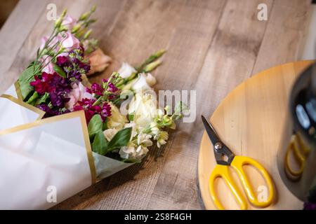 Splendido assortimento di fiori vivaci avvolti in carta con un paio di forbici con manici gialli nelle vicinanze su una superficie in legno, che raffigurano creatività e preparazione Foto Stock