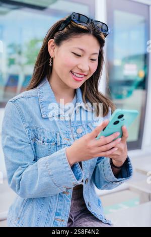 Giovane donna che usa lo smartphone alla stazione del tram con giacca in denim. Verticale Foto Stock