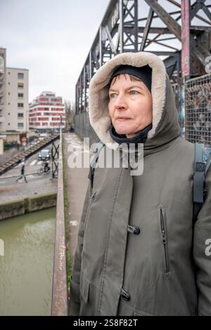 Una donna bianca matura che indossa una giacca con cappuccio kaki si erge su un ponte e guarda all'orizzonte in inverno Foto Stock