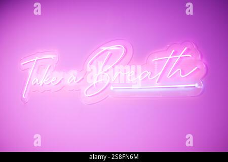 Un luccicante cartello al neon con le parole fai un respiro in corsivo, illuminato in rosa su uno sfondo coordinato, l'immagine evoca un'atmosfera rilassante e rilassante Foto Stock