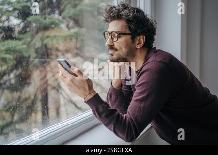 Un uomo medio-orientale adulto con gli occhiali sta scorrendo sul cellulare la mattina vicino alla finestra. Foto Stock
