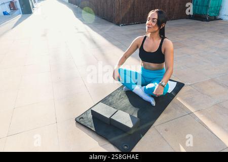 Una donna è seduta su un tappetino con un blocco di yoga davanti a lei. Indossa una canotta nera e pantaloni blu. La scena è tranquilla e serena, con Foto Stock