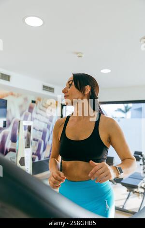 Una donna corre su un tapis roulant in una palestra. Indossa una canotta nera e pantaloni blu. Foto Stock