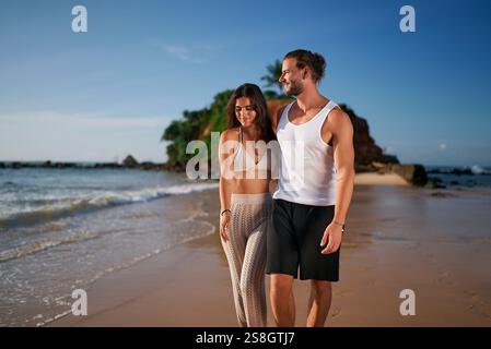 Una coppia di adulti gode di una tranquilla passeggiata sulla spiaggia al tramonto. Coppia spensierata senza bambini che passeggiano lungo il litorale. I partner romantici abbracciano il tempo libero e le vacanze al mare Foto Stock