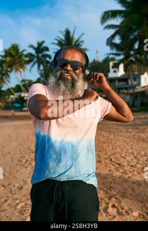 L'uomo con la barba, occhiali da sole, ama la musica con le cuffie wireless. Balla gioiosamente sulla soleggiata spiaggia tropicale. Buon nonno, look elegante, vivace Foto Stock
