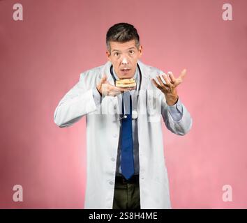 Giovane e affascinante medico con il viso sorpreso in abito, stetoscopio intorno al collo, cravatta e camicia, tenendo una torta e una mano con crema e un po' di crea Foto Stock