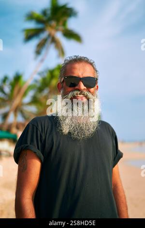 Uomo con barba anziana e occhiali da sole sulla spiaggia tropicale. Sorridendo in vacanza, cielo vibrante. Viaggi di piacere per uno stile di vita attivo. Divertimento all'aria aperta, avvolgente Foto Stock