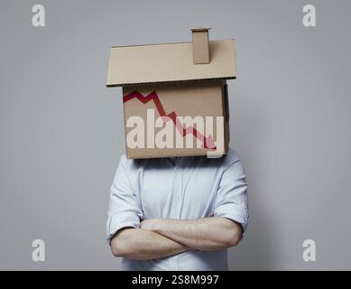 Uomo con una casa di cartone in testa e un grafico finanziario in declino con la freccia rivolta verso il basso, concetto di crisi del mercato immobiliare Foto Stock