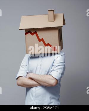Uomo con una casa di cartone in testa e un grafico finanziario in declino con la freccia rivolta verso il basso, concetto di crisi del mercato immobiliare Foto Stock