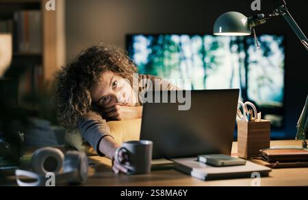 Una donna stanca e assonnata che lavora sul suo portatile a tarda notte, è seduta alla scrivania nel suo ufficio domestico e fissa lo schermo del computer Foto Stock