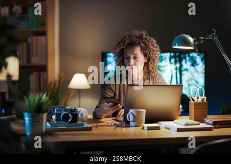 Giovane donna seduta alla scrivania nel suo ufficio domestico, sta lavorando sul suo computer portatile e sta controllando il suo smartphone Foto Stock