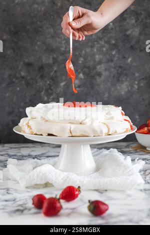 Una mano che aggiunge con cura la salsa di fragole su una pavlova ricoperta di panna montata, adagiata su un piedistallo bianco su una superficie di marmo. Foto Stock