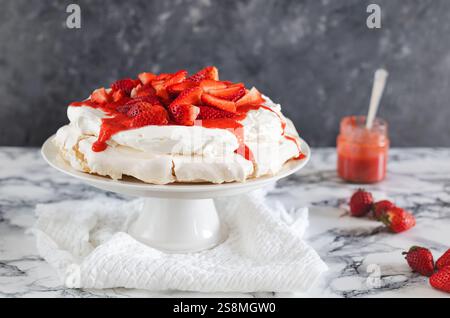 Una classica pavlova ricoperta da panna montata, fragole fresche e salsa di fragole su un supporto per torte bianco su una superficie di marmo Foto Stock