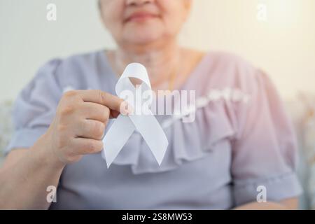 La messa a fuoco selettiva della mano femminile più anziana sta tenendo in mano un nastro bianco sullo sfondo di casa. Un simbolo della lotta contro il cancro ai polmoni. Novembre è il lattone polmonare Foto Stock