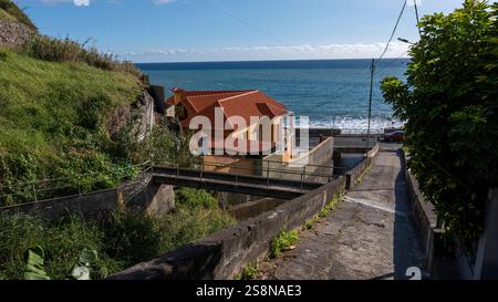 Una casa costiera con un tetto di piastrelle rosse, annidata tra lussureggianti colline verdi e l'oceano. Un sentiero in cemento in pendenza conduce al mare, creando un Foto Stock