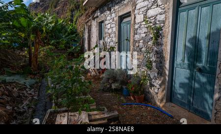 Una casa rustica in pietra con porte in legno blu intemprato, circondata da lussureggianti piante di banane e vegetazione in vaso. L'affascinante ambiente rurale riflette una favola Foto Stock