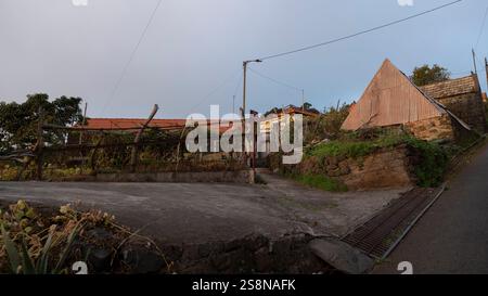 Un villaggio rurale caratterizzato da vecchie case, una recinzione in legno e una struttura dal tetto triangolare su una collina. L'ambiente rustico riflette la tradizione Foto Stock