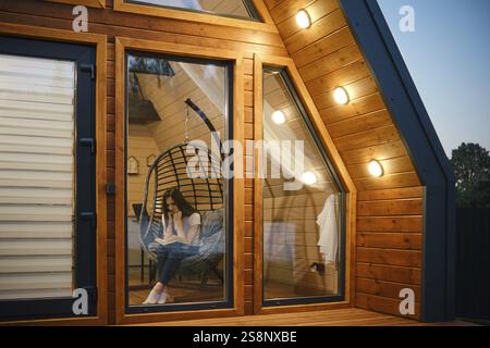 Donna che si rilassa su una sedia sospesa con un libro all'interno di un'elegante cabina in legno. La calda illuminazione e le grandi finestre creano un'atmosfera invitante, perfec Foto Stock