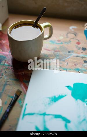 La tazza da caffè si siede su un tavolo colorato e ricoperto di vernice accanto a una tela con pennellate astratte color acqua, suggerendo una pausa nel lavoro di un artista Foto Stock