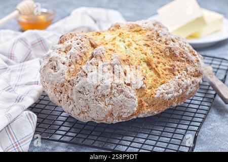 Una pagnotta rustica di pane sodato poggia su una griglia di raffreddamento. Il burro si siede su un piatto nelle vicinanze, accompagnato da un barattolo di miele, creando un'atmosfera accogliente in cucina, orizzontale Foto Stock