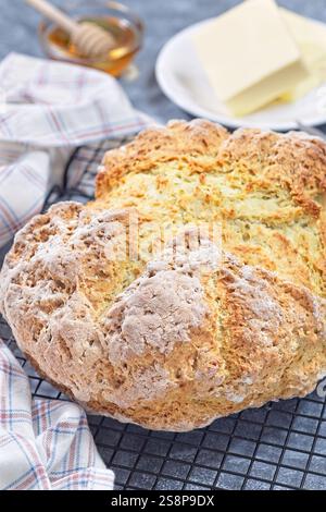 Una pagnotta rustica di pane sodato poggia su una griglia di raffreddamento. Il burro si siede su un piatto nelle vicinanze, accompagnato da un barattolo di miele, creando un'atmosfera accogliente da cucina e un primo piano verticale Foto Stock