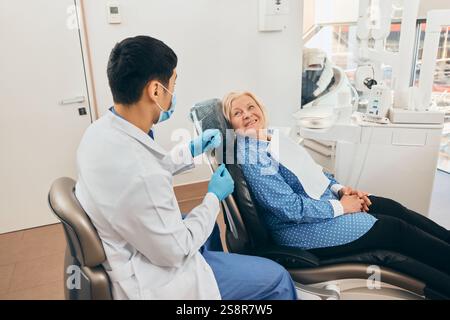 Medico maschile in maschera e guanti che parla con una donna anziana sorridente sdraiata rilassata in poltrona dentale, moderni strumenti dentali visibili in ambiente clinico. Foto Stock