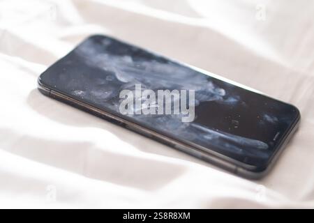 Primo piano di uno smartphone adagiato su una superficie morbida, che mostra uno schermo macchiato con impronte digitali e polvere. Lo sfondo è leggermente sfocato, enfasi Foto Stock