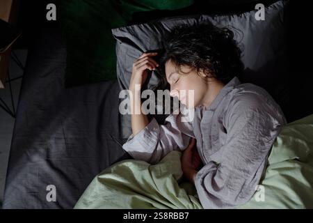 Colpo dall'alto di donna bruna dai capelli ricci vestita con pigiami di cotone che si gode di dormire in un letto nuovo con biancheria di cotone Foto Stock