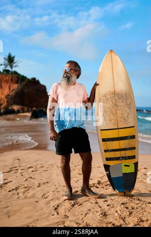 L'uomo anziano con tavola da surf si trova sulla spiaggia sabbiosa. Gli anziani surfisti amano le giornate di sole sull'oceano. Stile di vita attivo per gli adulti più anziani. Onde marine, cielo blu dentro Foto Stock