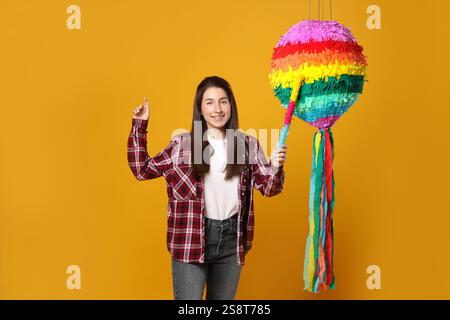 Donna felice che colpisce pinata colorata con bastoncino su sfondo arancione Foto Stock