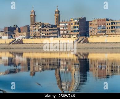 Una tranquilla cittadina lungo il fiume di Naqadah in Egitto, con edifici tradizionali e vegetazione lussureggiante, con deserto del Sahara alle spalle Foto Stock