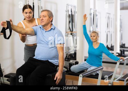 Giovane istruttrice positiva che controlla i movimenti dell'uomo anziano che si allunga sulle attrezzature del riformatore Pilates in studio Foto Stock