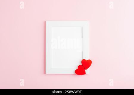 Cornice fotografica bianca con due cuori rossi su sfondo rosa chiaro. Vista dall'alto, posizione piatta, mockup. Foto Stock