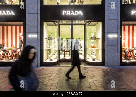 Lugano, Svizzera - 22 dicembre 2024: Persone che camminano davanti a un negozio illuminato di Prada la sera Foto Stock