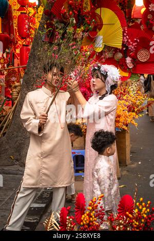 Una giovane famiglia vestita con costumi tradizionali ao dai e con un hoa dao, o un ramo in fiore di pesca, posare per le foto a un Tet o al mercato lunare di Capodanno, Foto Stock