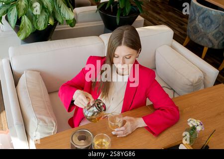 Donna con una giacca rossa che versa il tè in una tazza. La scena è ambientata in un'atmosfera intima e intima, con un tavolo e sedie in legno. La donna sta godendo Foto Stock