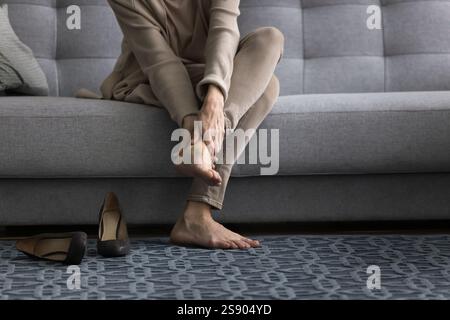 Donna di mezza età seduta sul divano a piedi nudi massaggiando i piedi affaticati Foto Stock