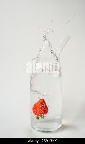 Spruzzare fragole in un bicchiere d'acqua con cannuccia riutilizzabile su bianco Foto Stock