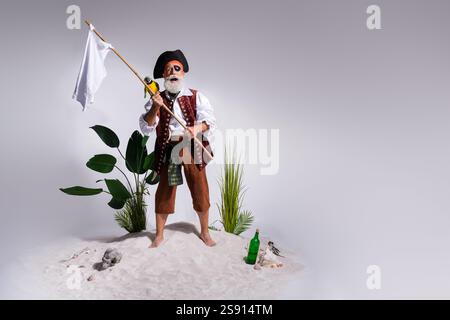 Un anziano pirata barbuto in costume da bucaniere si erge su una spiaggia di sabbia bianca che regge bandiera, ed esotiche spedizioni sull'isola. Foto Stock